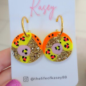 May include: A pair of vibrant hoop earrings. Each earring has a circular design with yellow, orange, and gold glitter elements. The design includes pink and red leopard print details. The earrings are attached to gold hoops.