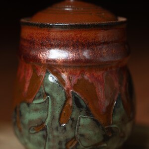 May include: A handcrafted ceramic jar with a lid. The jar features a rich color palette of brown, red, and green, with a textured surface and leaf-like patterns. The jar is approximately 5 inches tall.