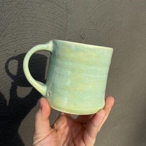 May include: A handmade ceramic mug with a light green glaze. The mug has a rounded body, a curved handle, and a small star-shaped detail near the base of the handle. The mug is held in a hand against a dark background.