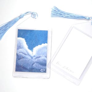 May include: Two rectangular clear plastic bookmarks with light blue tassels. One bookmark features a small painting of white clouds against a deep blue sky with a signature in the corner. The other bookmark is blank with faint handwritten text.