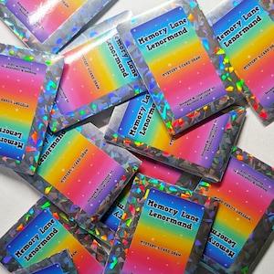 May include: A collection of holographic card packs with the text "Memory Lane Lenormand" and "Mystery 3-Card Draw." Each pack features a rainbow gradient design and is designed and illustrated by Rainbow Willard.