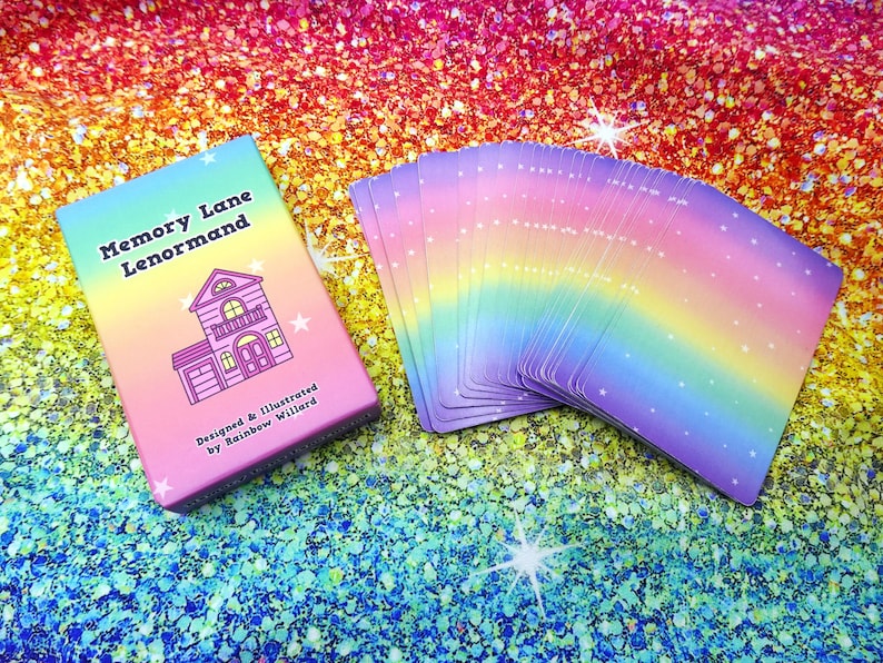 May include: A deck of Memory Lane Lenormand cards with a matching box. The box is pink and features a house illustration and the text "Memory Lane Lenormand". The cards have a rainbow gradient design with white stars.
