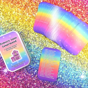 May include: A Memory Lane Lenormand divination deck in a metal tin, a stack of rainbow-gradient cards with white stars, and a reference card listing meanings. The tin features a house illustration and text: Memory Lane Lenormand, A Nostalgic 30-Card Divination Deck, Designed & Illustrated by Rainbow Willard.