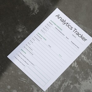 May include: A white paper titled "Analytics Tracker" on a gray surface. The paper includes sections for platform overview, monthly stats, and insights. The text is in black and green, with lines for filling in data. The paper is angled in the image.