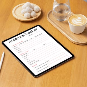 May include: A digital tablet displaying an "Analytics Tracker" form, alongside a white stylus, a cup of coffee, a glass of water, and a small plate of white confections, all arranged on a wooden table. The tablet's screen features a detailed layout for tracking various metrics.