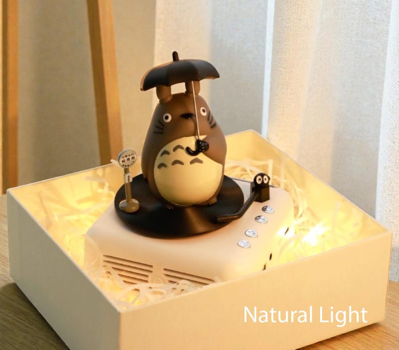 Pu&ograve; includere: Una statuetta decorativa di Totoro con un ombrello, sopra un altoparlante bianco con un braccio di giradischi nero. L'altoparlante &egrave; all'interno di una scatola bianca con luci fatate. Il testo "Natural Light" &egrave; visibile.
