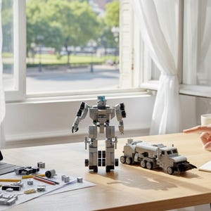 May include: A light grey robot and a matching truck model made of interlocking blocks sit on a wooden table. Various blocks and tools are scattered around, with a tablet displaying a diagram. A hand points towards the truck.