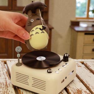 Pu&ograve; includere: Un giradischi in stile retr&ograve; color crema con un disco in vinile nero e una statuetta di un personaggio dei cartoni animati. La statuetta &egrave; marrone con la pancia gialla e tiene un piccolo ombrello. Il giradischi ha una piccola antenna e una porta USB.