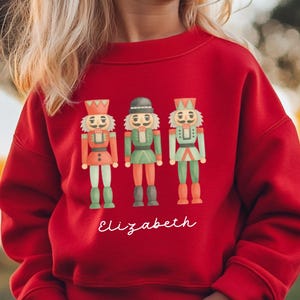 May include: A bright red long-sleeve sweatshirt featuring a graphic print of three nutcracker figures in red, green, and light green uniforms. Below the nutcrackers, the name Elizabeth is printed in a white, cursive script font.