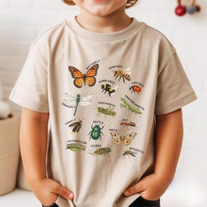 May include: A beige short-sleeved t-shirt featuring a detailed graphic print of various insects and their names, including a butterfly, honeybee, bumblebee, ladybug, dragonfly, grasshopper, praying mantis, firefly, beetle, ant, caterpillar, cicada, moth, and mosquito.