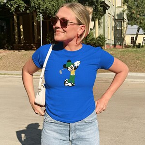 May include: A woman wearing a royal blue t-shirt featuring a cartoon Mickey Mouse character playing golf. The shirt is paired with light-wash denim shorts and a white shoulder bag. The person is also wearing sunglasses.