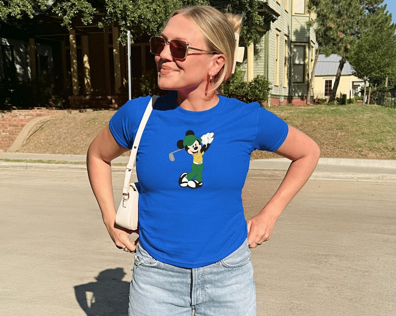 May include: A woman wearing a royal blue t-shirt featuring a cartoon Mickey Mouse character playing golf. The shirt is paired with light-wash denim shorts and a white shoulder bag. The person is also wearing sunglasses.