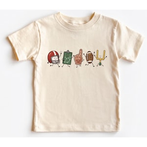 May include: A cream-colored short-sleeve t-shirt featuring a graphic print of five animated American football-themed characters: a red helmet, a green play clipboard, a brown foam finger, a brown football, and a yellow goal post.