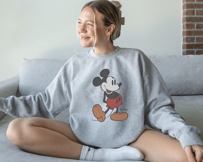 May include: A light gray sweatshirt featuring a classic Mickey Mouse graphic. The cartoon character is depicted in his iconic red shorts, brown shoes, and black gloves. The sweatshirt has a crew neck and long sleeves.
