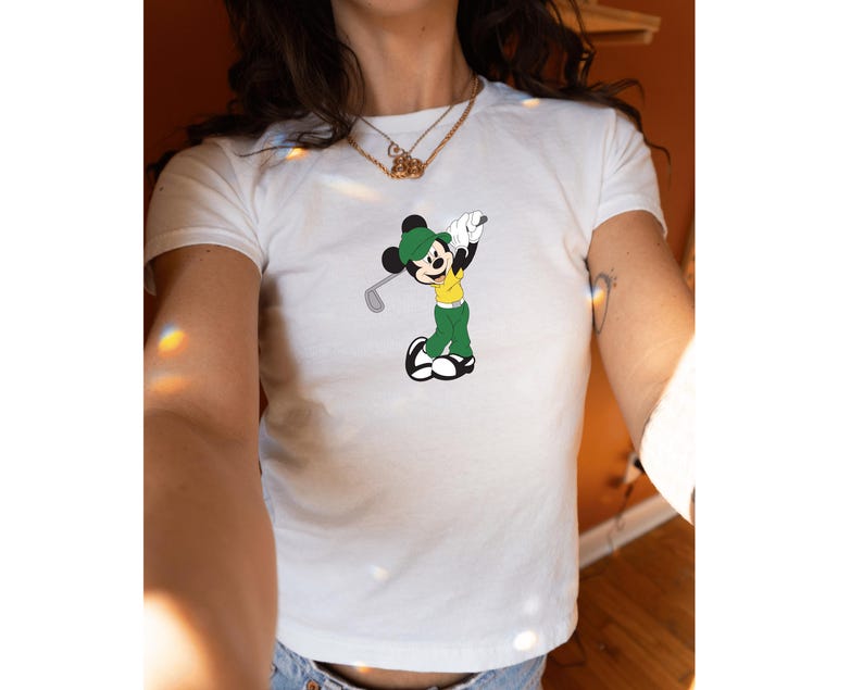 May include: White t-shirt featuring a cartoon Mickey Mouse character dressed in golf attire. Mickey is wearing a green hat, green pants, a yellow shirt, and black shoes, holding a golf club. The design is centered on the shirt.