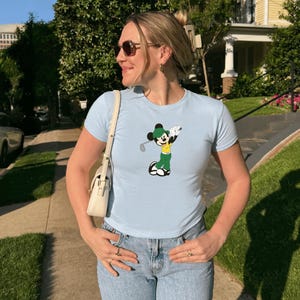 May include: Light blue t-shirt featuring a cartoon Mickey Mouse playing golf. The graphic shows Mickey in a green outfit with a golf club. The shirt is paired with light wash jeans and a white purse.