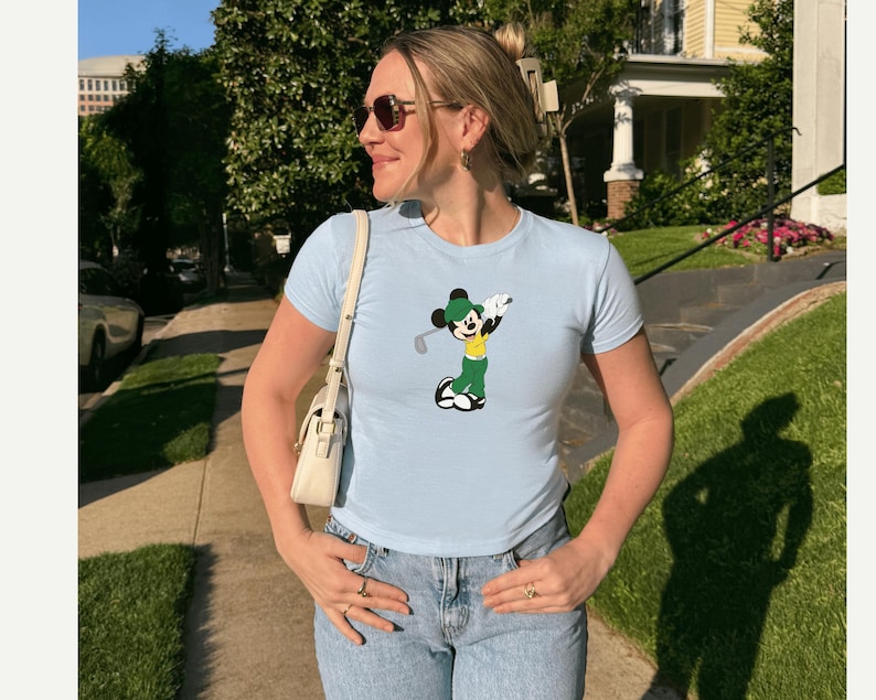 May include: Light blue t-shirt featuring a cartoon Mickey Mouse playing golf. The graphic shows Mickey in a green outfit with a golf club. The shirt is paired with light wash jeans and a white purse.