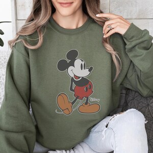 以下が含まれることがあります： ミッキーマウスのグラフィックが特徴のオリーブグリーンのスウェットシャツ。漫画のキャラクターは、赤いショートパンツ、茶色の靴、黒い耳を身に着けて、彼の象徴的なポーズで描かれています。ディズニーのロゴが右下に印刷されています。