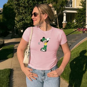 May include: A light pink t-shirt featuring a cartoon Mickey Mouse character dressed in golf attire. The character is depicted in green and yellow, holding a golf club. The shirt is short-sleeved and worn with blue jeans.