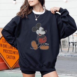 May include: Navy blue sweatshirt featuring a classic Mickey Mouse graphic. The cartoon character is depicted in his iconic pose, wearing red shorts and brown shoes. The sweatshirt is a comfortable, casual style.