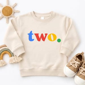 May include: A beige long-sleeve crewneck sweatshirt featuring the word "two." printed in the center. The letter "t" is blue, "w" is red, and "o" is yellow, followed by a small green period. The sweatshirt has ribbed cuffs and a ribbed neckline.