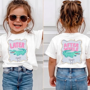 May include: A white short-sleeve t-shirt featuring a graphic print of a green alligator inside a blue and white striped frame. The front displays the pink text LATER, while the back displays the pink text AFTER WHILE, both with a bow icon.