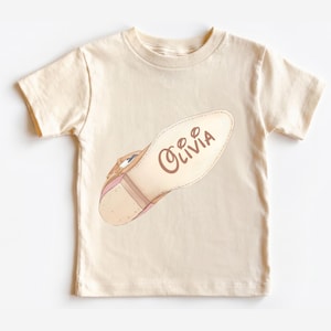 May include: A cream-colored short-sleeve t-shirt featuring a graphic of a tan shoe sole on the front. The sole has a brown buckle detail at the heel and the name Olivia written in a brown cursive script across the center of the shoe sole.