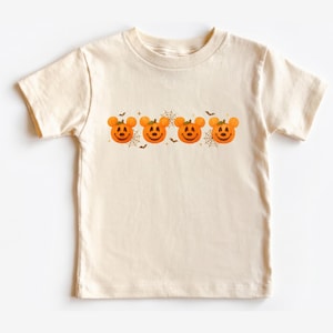 May include: A cream-colored short-sleeve t-shirt featuring a horizontal graphic print of four orange jack-o'-lanterns with round ears, interspersed with thin brown spider web illustrations and small flying bat silhouettes across the chest.