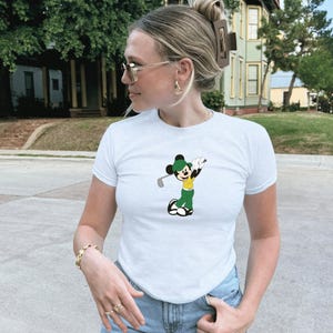 May include: White short-sleeve t-shirt featuring a cartoon Mickey Mouse character in a golf pose. The character is wearing a green hat, yellow shirt, and green pants. The shirt is a classic crew neck style.