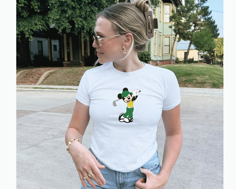 May include: White short-sleeve t-shirt featuring a cartoon Mickey Mouse character in a golf pose. The character is wearing a green hat, yellow shirt, and green pants. The shirt is a classic crew neck style.