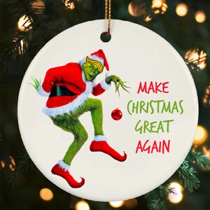 May include: Round Christmas ornament featuring a green character in a red Santa suit with the text "MAKE CHRISTMAS GREAT AGAIN" in green and red. The ornament has a gold string for hanging.