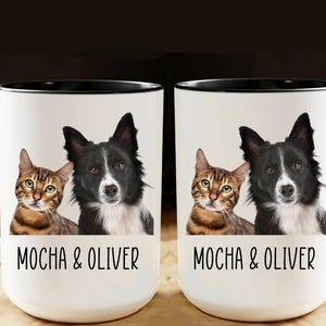 May include: White ceramic mug with a black handle and rim, featuring a cat and dog portrait. The cat is a brown tabby, and the dog is a black and white Border Collie. The mug has the text "MOCHA & OLIVER" in black.