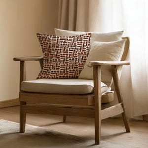 May include: A wooden armchair with a beige cushion and pillows. The patterned pillow features a repeating design in shades of brown and red on a white background. The chair is positioned in front of a neutral-colored curtain.