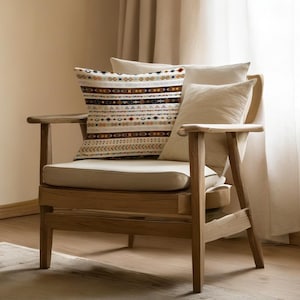 May include: A wooden armchair with a beige cushion and backrest. A decorative pillow with a geometric pattern in brown, beige, and blue tones rests on the chair, along with two solid beige pillows. The chair is positioned near a window with beige curtains.