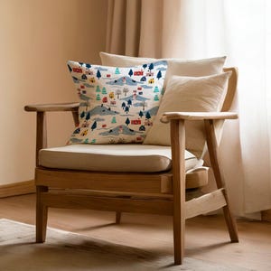 Puede incluir: Un sillón de madera con un cojín y respaldo color crema. Una almohada decorativa con un estampado de escena de pueblo con casas, árboles y colinas se coloca en la silla. Una almohada más pequeña y lisa de color crema descansa contra el respaldo.