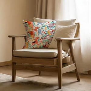 May include: A decorative throw pillow with a whimsical pattern of colorful animals and floral elements. The pillow features a cream-colored background with illustrations of llamas, elephants, and other creatures. The pillow is placed on a wooden chair with cream-colored cushions.