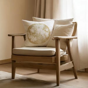 Può includere: Una poltrona in legno con cuscino e schienale color crema. La sedia ha una struttura e braccioli marrone chiaro. Un cuscino decorativo con un disegno di luna dorata è sulla sedia, insieme ad altri cuscini di colore neutro. La sedia è davanti a una finestra con tende trasparenti.