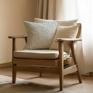 Puede incluir: Un sillón de madera con un cojín y respaldo de color beige claro. La silla tiene una almohada con estampado floral en tonos azules, naranjas y amarillos. La silla está colocada frente a una cortina beige.