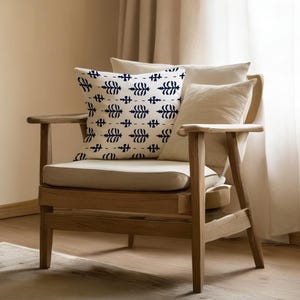 May include: A wooden armchair with a cream cushion and backrest. A decorative pillow with a navy and white pattern sits on the chair. Additional cream pillows provide support. The chair is in front of a neutral-coloured curtain.