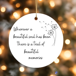 May include: A white, round Christmas ornament with a gold string. The ornament features black text that reads "Wherever a beautiful soul has been there is a trail of beautiful memories." A black and white dandelion design is also present.