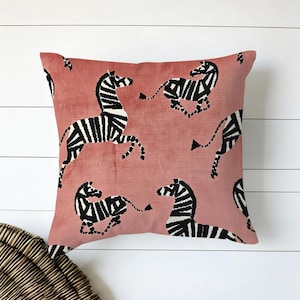 May include: A square cushion with a dusty rose hue, adorned with a repeating pattern of black and white zebras in different poses. The cushion has a soft, textured look and is set against a white wooden backdrop.