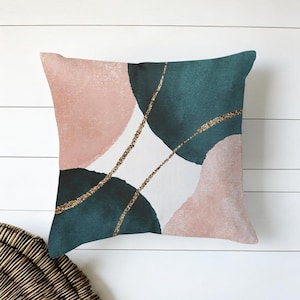 Puede incluir: Cojín decorativo cuadrado con un diseño abstracto. El cojín presenta formas de acuarela verde azulado y rosa rubor con detalles de purpurina dorada. El cojín está sobre un fondo blanco.