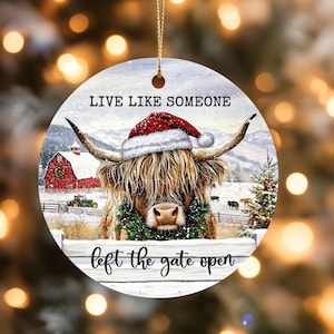 以下が含まれることがあります： サンタ帽とリースをつけたハイランド牛が描かれた丸いクリスマスオーナメント。「Live Like Someone」と「left the gate open」の文字が入っています。背景には、赤い納屋がある雪景色が描かれています。