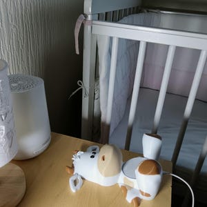 May include: A white and brown cow-shaped baby sound machine sits on a wooden surface. A white cot with a pink ribbon is in the background. A white lamp is on the left.