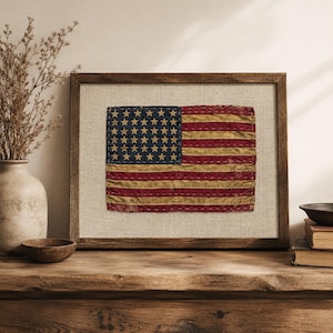 May include: A framed textile art piece featuring a weathered American flag with a blue canton containing stars and horizontal red and tan stripes. The flag is stitched onto a light beige fabric background and enclosed in a dark wooden frame.