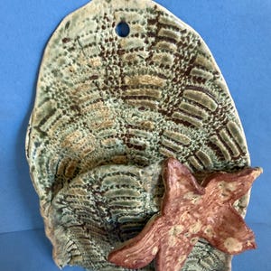 May include: A wall-mounted ceramic pocket with a textured, mottled green and brown finish. The piece features a prominent, raised, reddish-brown starfish attached to the front surface. A small circular hole is positioned at the top for hanging.