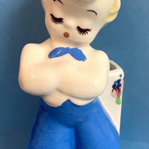 May include: A ceramic planter shaped like a boy with blue pants, a white shirt, and a blue bow tie. The planter has a white background with a floral design on the side.