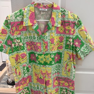 May include: A short-sleeved button-down shirt featuring a vibrant patchwork pattern of pink, yellow, and green floral and botanical motifs. The shirt has a classic collar, a front button closure, and a label at the neck that reads Sanibel.