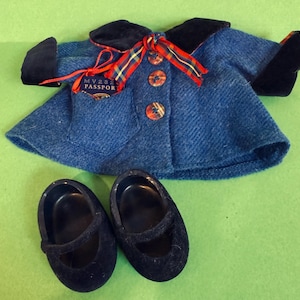 May include: A blue doll coat with black velvet trim, a red and gold plaid ribbon, and three decorative buttons. Two black doll shoes with a strap are also included. The coat has a small pocket with a tag that reads "MV232 PASSPORT".