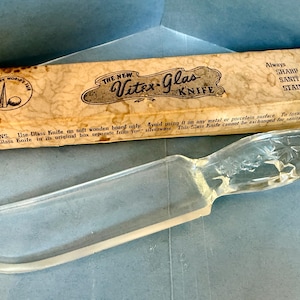 May include: A clear glass knife with a textured handle featuring star patterns, accompanied by its original rectangular cardboard box. The box is printed with "The New Vitex-Glas Knife," "New York World's Fair," and "Always Sharp Sanitary Stainless."